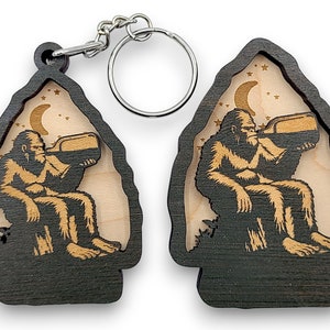 May include: A wooden keychain with a black and brown finish. The keychain features a silhouette of a Bigfoot sitting on a rock and drinking from a bottle under a crescent moon and stars.