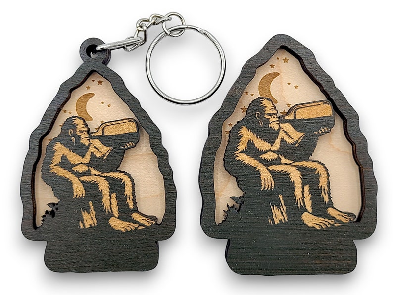 May include: A wooden keychain with a black and brown finish. The keychain features a silhouette of a Bigfoot sitting on a rock and drinking from a bottle under a crescent moon and stars.