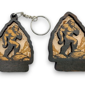 May include: A wooden keychain featuring a silhouette of Bigfoot walking through a forest scene. The keychain is dark brown with a light brown interior and a silver metal ring.