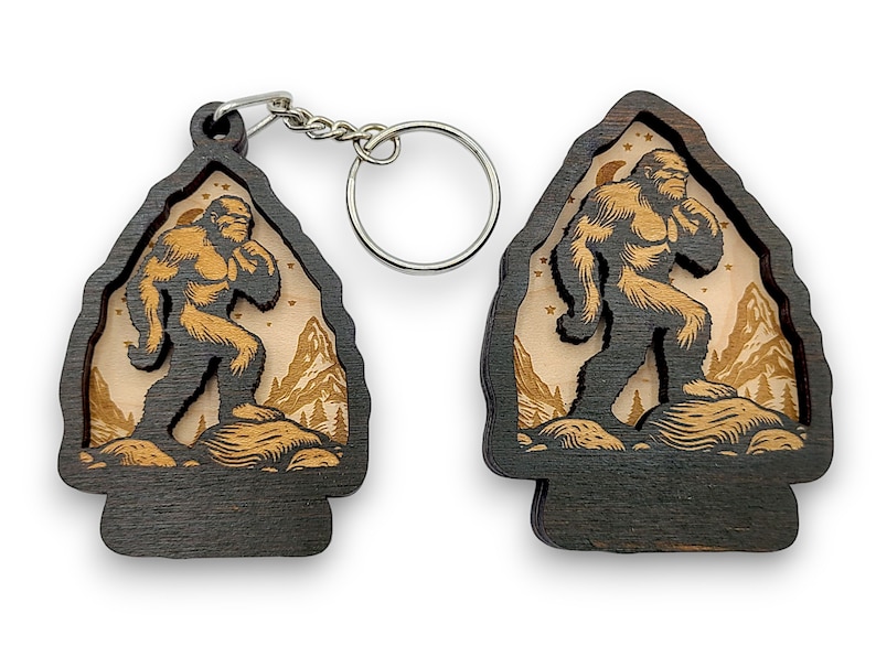 May include: A wooden keychain featuring a silhouette of Bigfoot walking through a forest scene. The keychain is dark brown with a light brown interior and a silver metal ring.