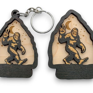 May include: A wooden keychain featuring a Bigfoot silhouette with a peace sign, walking under a crescent moon and stars. The keychain is made of dark wood with a light wood insert.
