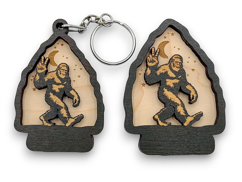 May include: A wooden keychain featuring a Bigfoot silhouette with a peace sign, walking under a crescent moon and stars. The keychain is made of dark wood with a light wood insert.