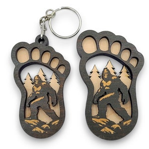May include: A wooden keychain shaped like a Bigfoot footprint with a detailed image of Bigfoot walking through a forest inside the footprint.