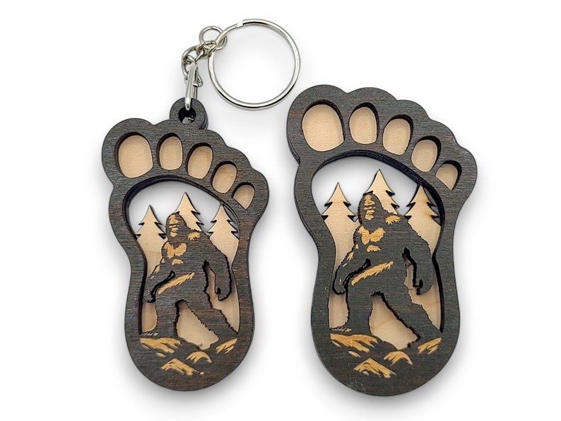 May include: A wooden keychain shaped like a Bigfoot footprint with a detailed image of Bigfoot walking through a forest inside the footprint.