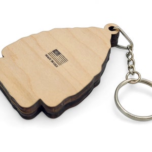 May include: Wooden keychain shaped like a Christmas tree with a brown wood grain finish. The back of the keychain has the text "Made in USA" with a small American flag graphic.