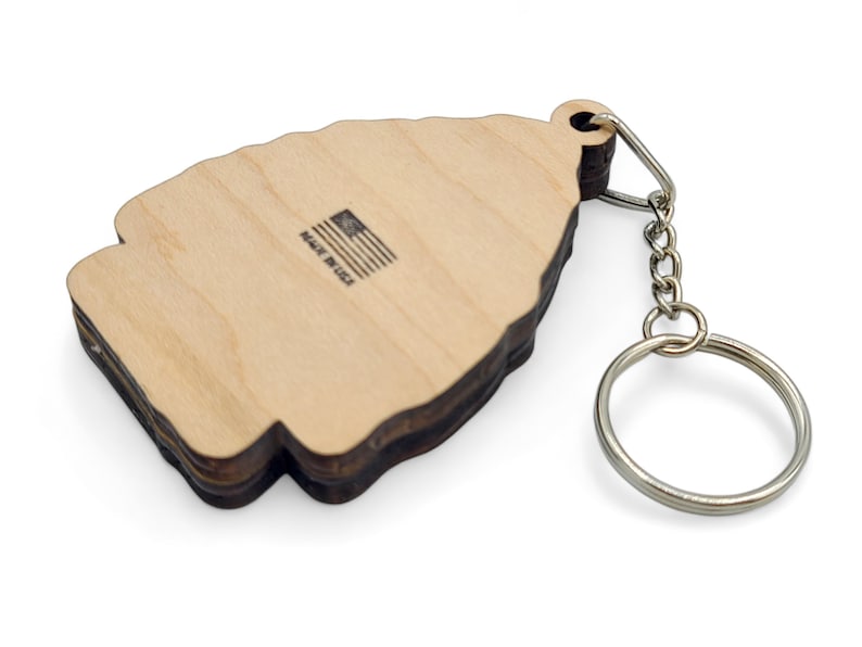 May include: Wooden keychain shaped like a Christmas tree with a brown wood grain finish. The back of the keychain has the text "Made in USA" with a small American flag graphic.