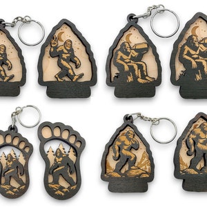 May include: Six different wooden keychains featuring a Bigfoot design. The keychains are in the shapes of a footprint, a mountain, and an arrowhead. The Bigfoot is depicted in different poses, including walking, sitting, and drinking. The keychains are made of dark wood with a light wood inlay.