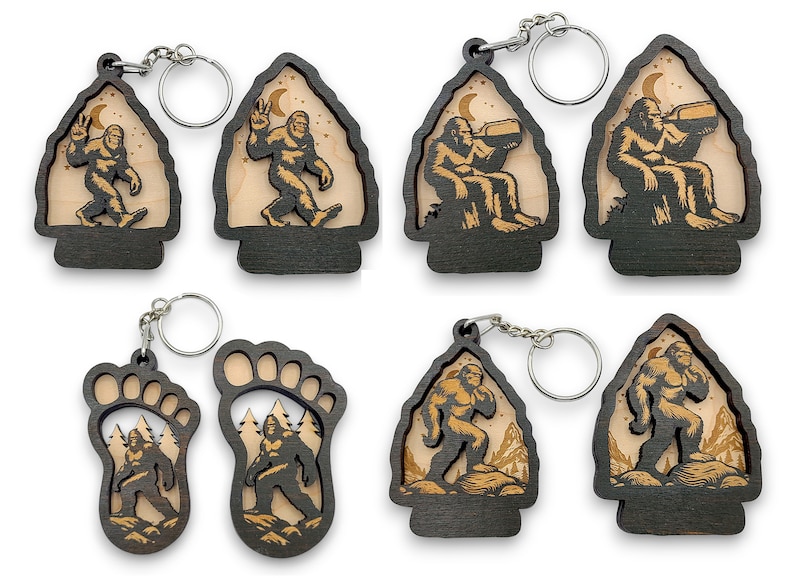 May include: Six different wooden keychains featuring a Bigfoot design. The keychains are in the shapes of a footprint, a mountain, and an arrowhead. The Bigfoot is depicted in different poses, including walking, sitting, and drinking. The keychains are made of dark wood with a light wood inlay.