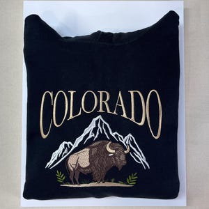May include: A black hooded sweatshirt featuring an embroidered design on the front. The text "COLORADO" is stitched in a light tan color above a white mountain range graphic. Below the mountains is a brown bison with small green leaf accents.