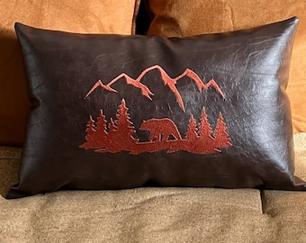 May include: A brown faux leather pillow with a red embroidered design of mountains, trees, and a bear.