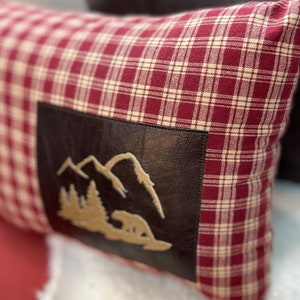 May include: Red and white plaid pillow with a brown leather patch featuring an embroidered mountain scene.