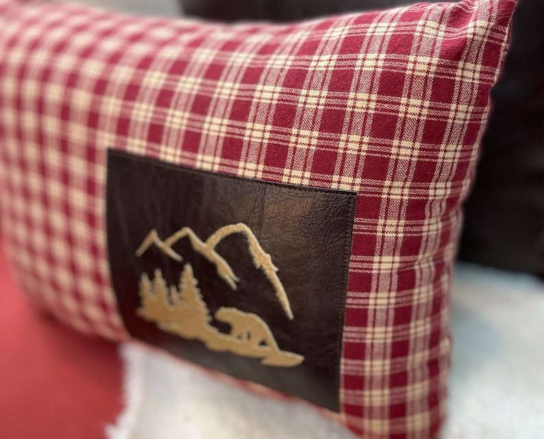 May include: Red and white plaid pillow with a brown leather patch featuring an embroidered mountain scene.