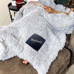 May include: A soft, light gray faux fur blanket with a black square patch that reads "Weekends" in cursive. The blanket is draped over a brown armchair, creating a cozy and inviting scene. The blanket's texture is fluffy and shaggy.