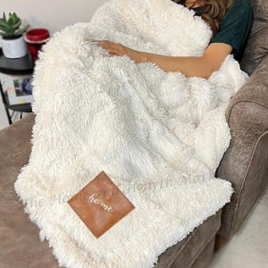 May include: A cream-colored, fluffy blanket with a brown square patch that reads "home" in white script. The blanket is draped over a person seated in a brown armchair. The blanket has a soft, textured appearance.