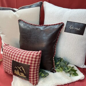 May include: Four decorative throw pillows on a white faux fur throw. The pillows are in various colors and textures, including red, white, brown, and black. One pillow has the word "home" embroidered on it.