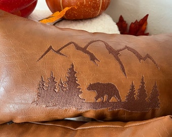 May include: Brown rectangular pillow with an embroidered mountain and bear scene. The pillow has a leather-like texture and a zipper closure. The design features a bear walking in front of a forest and mountains.