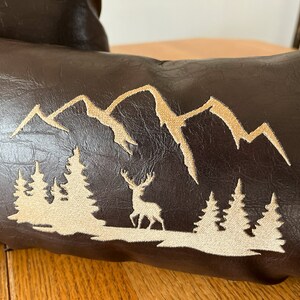 May include: A brown faux leather pillow with an embroidered design of a mountain range, trees, and a deer. The embroidery is in a light beige color.