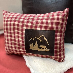 May include: Red and white plaid throw pillow with a brown leather patch featuring a mountain scene and a bear silhouette.