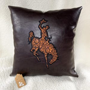 May include: Brown leather pillow with a tooled leather design of a bucking bronco. The pillow has the text "The Home and Heart" on the bottom.