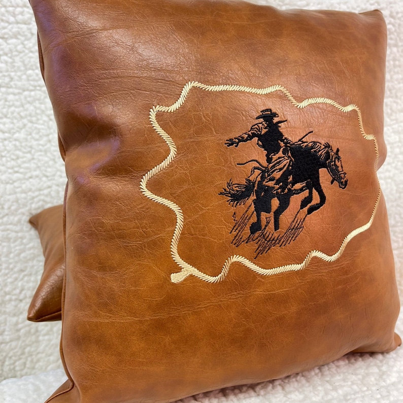 May include: A brown leather pillow with a stitched design of a cowboy riding a horse. The design is in black thread and is surrounded by a white stitched border.