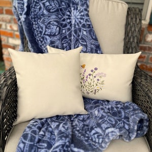 Pode incluir: Duas almofadas de cor creme com desenhos florais bordados est&atilde;o em uma cadeira de vime com um cobertor azul e branco.