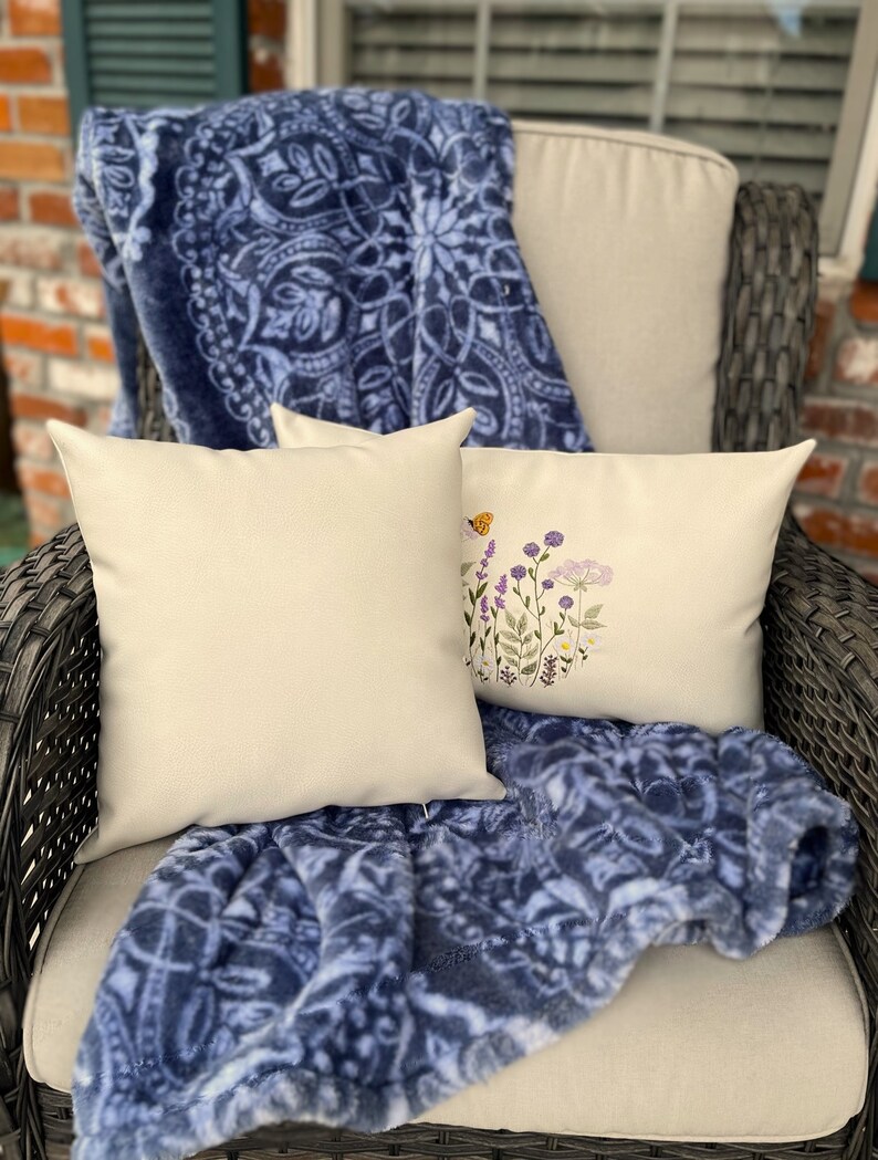 Pode incluir: Duas almofadas de cor creme com desenhos florais bordados est&atilde;o em uma cadeira de vime com um cobertor azul e branco.