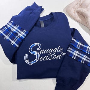 May include: A navy blue crewneck sweatshirt featuring the white text Snuggle Season with a small heart icon. The sleeves are accented with two horizontal bands of blue and white plaid fabric that have frayed, raw edges for a textured look.