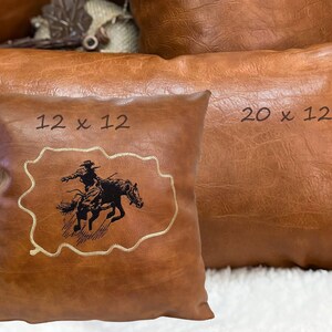 May include: Two brown leather pillows with embroidered designs. The smaller pillow is 12 x 12 inches and features a cowboy riding a bucking horse. The larger pillow is 20 x 12 inches.