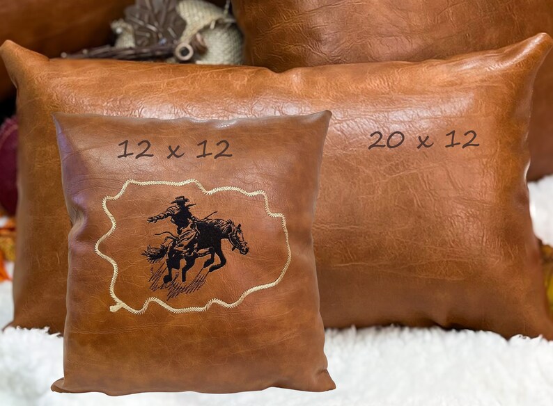 May include: Two brown leather pillows with embroidered designs. The smaller pillow is 12 x 12 inches and features a cowboy riding a bucking horse. The larger pillow is 20 x 12 inches.