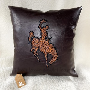 May include: Brown leather pillow with a tooled leather design of a bucking bronco. The pillow has the text "The Home and Heart" on the bottom.