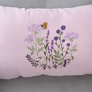 Pode incluir: Uma almofada rosa com flores silvestres bordadas e uma borboleta em tons de roxo, verde e amarelo.