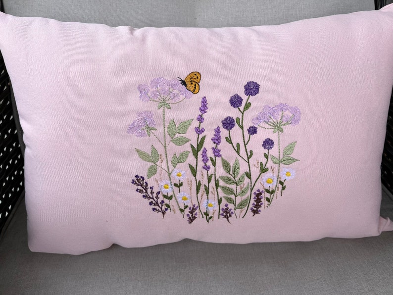 Pode incluir: Uma almofada rosa com flores silvestres bordadas e uma borboleta em tons de roxo, verde e amarelo.