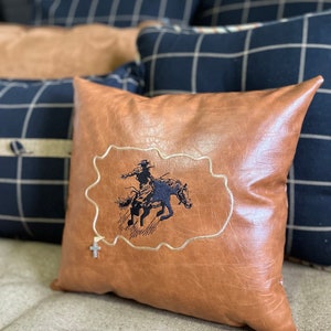 May include: A brown leather pillow with a black embroidered design of a cowboy riding a horse. The pillow has a silver cross charm attached to it.