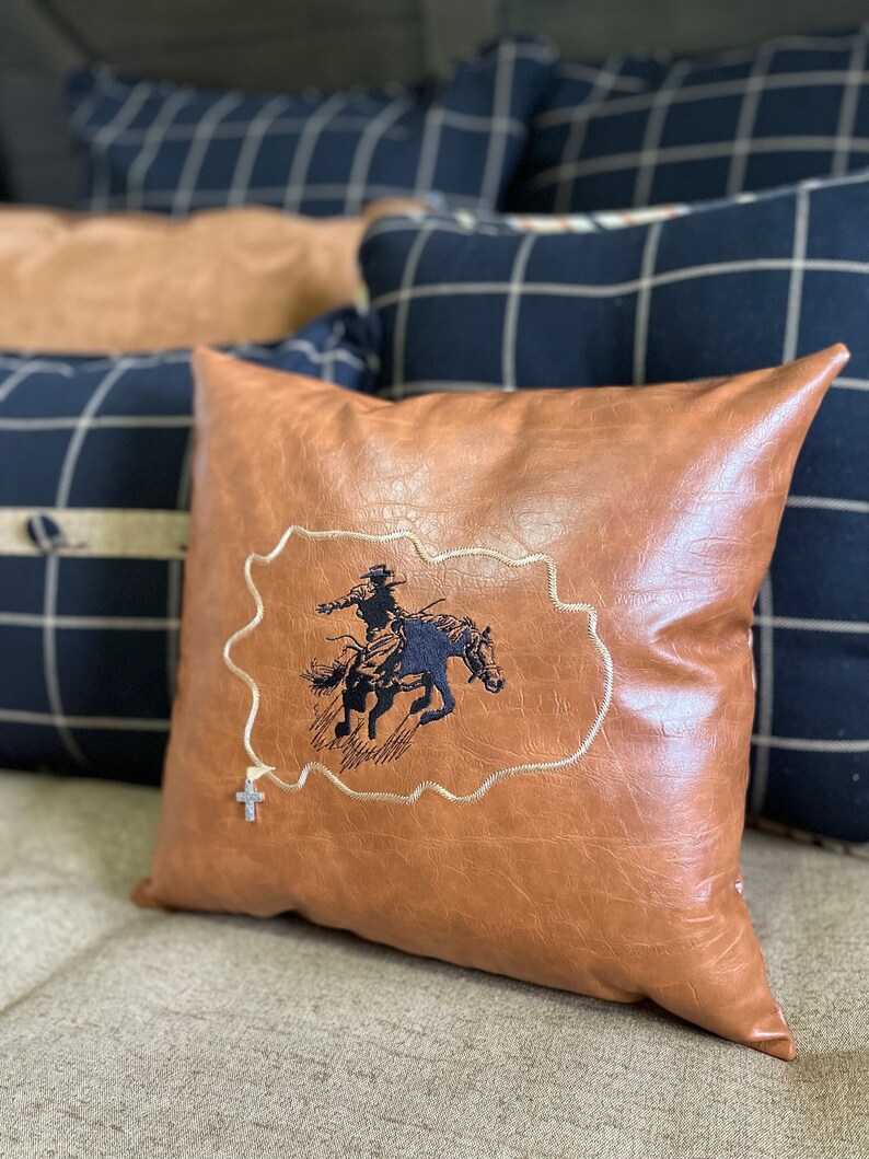 May include: A brown leather pillow with a black embroidered design of a cowboy riding a horse. The pillow has a silver cross charm attached to it.