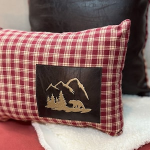 May include: Red and white plaid throw pillow with a brown leather patch featuring an embroidered design of mountains and a bear.