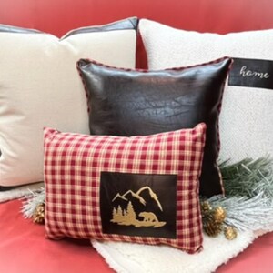 May include: Four decorative throw pillows with different patterns and textures. The pillows are arranged on a red surface with a white faux fur throw. The pillows include a white pillow with black trim, a brown leather pillow, a white pillow with the word "home" in black, and a red and white plaid pillow with a brown leather patch featuring a mountain scene and a bear.