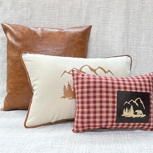 May include: Three decorative throw pillows. The largest pillow is brown faux leather. The middle pillow is white with a brown mountain and tree design. The smallest pillow is red and white plaid with a brown patch featuring a mountain and tree design with a bear.