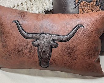 Può includere: Un cuscino rettangolare in similpelle marrone con una silhouette centrale di testa di toro longhorn marrone scuro. La testa del toro presenta un intricato motivo floreale in rilievo ed è delineata da cuciture nere sullo sfondo marrone testurizzato.