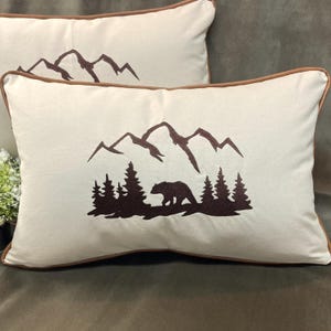 May include: Two rectangular throw pillows with a cream-colored background. Each pillow features a brown mountain range design, a bear, and trees. The pillows have brown piping around the edges, adding a decorative touch.