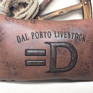 May include: A rectangular brown pillow with the text "DAL PORTO LIVESTOCK" and a large letter "D" with two horizontal bars. The pillow has a textured, leather-like appearance. The background includes a rope and a wooden wheel.