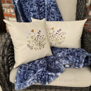 Pode incluir: Duas almofadas brancas com desenhos florais bordados e borboletas. As almofadas est&atilde;o sobre uma manta de l&atilde; azul e branca com padr&otilde;es e uma cadeira de vime.