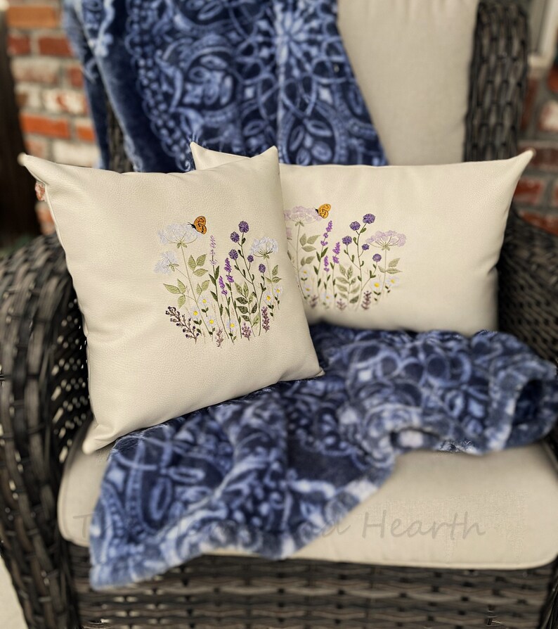 Pode incluir: Duas almofadas brancas com desenhos florais bordados e borboletas. As almofadas est&atilde;o sobre uma manta de l&atilde; azul e branca com padr&otilde;es e uma cadeira de vime.