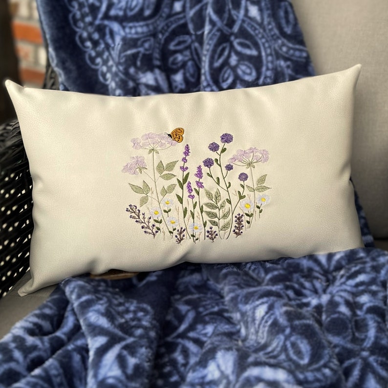Pode incluir: Uma almofada branca com flores roxas e brancas bordadas e uma pequena borboleta laranja. A almofada est&aacute; sobre uma manta de tecido azul e branca com padr&atilde;o.