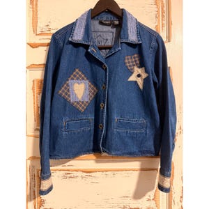 May include: A blue denim jacket featuring a collared neckline with light blue textured trim. The front displays a diamond-shaped patch with a heart, a plaid heart patch, and a star-shaped patch with a button. It has long sleeves with plaid cuffs.