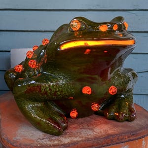 May include: A dark green ceramic frog figurine featuring a glossy finish and multiple embedded orange glass accents that glow from within. The frog has a wide mouth and textured skin, with light emanating from its eyes, mouth, and various spots on its back.