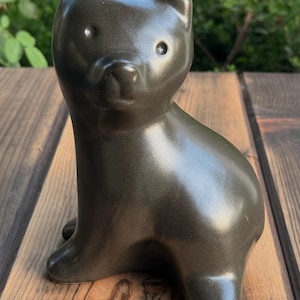 Könnte beinhalten: Eine glatte, dunkel anthrazitfarbene Bärenfigur mit glänzender Oberfläche. Der Bär ist in sitzender Position dargestellt und blickt nach vorne, mit kleinen, abgerundeten Ohren, einfachen kreisförmigen Augen und einer ausgeprägten Schnauze.