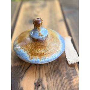 May include: A round ceramic oil lamp with a central chimney, featuring a mottled glaze in shades of brown, tan, and light blue. The lamp has a smooth, rounded body and a distinct top opening. A white woven cotton wick lies beside the lamp.