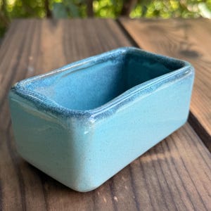 May include: A rectangular ceramic vessel with a smooth, glossy finish in a vibrant turquoise blue colour. The container features rounded corners and a slightly darker, mottled blue glaze along the top rim, creating a subtle contrast in texture.