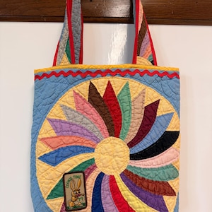 May include: A quilted tote bag featuring a light blue background and a central sunburst pattern made of multi-colored fabric petals. A rectangular patch with a cartoon rabbit and the text "COOKIE TIME" is sewn onto the lower left side.