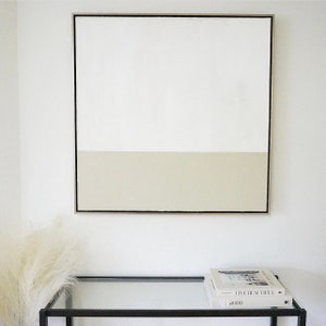 May include: A framed abstract painting with a white top half and a light beige bottom half. The painting is hanging on a white wall above a glass-top table with two books stacked on top of it. The books are titled "Live Beautiful" and "at 100".